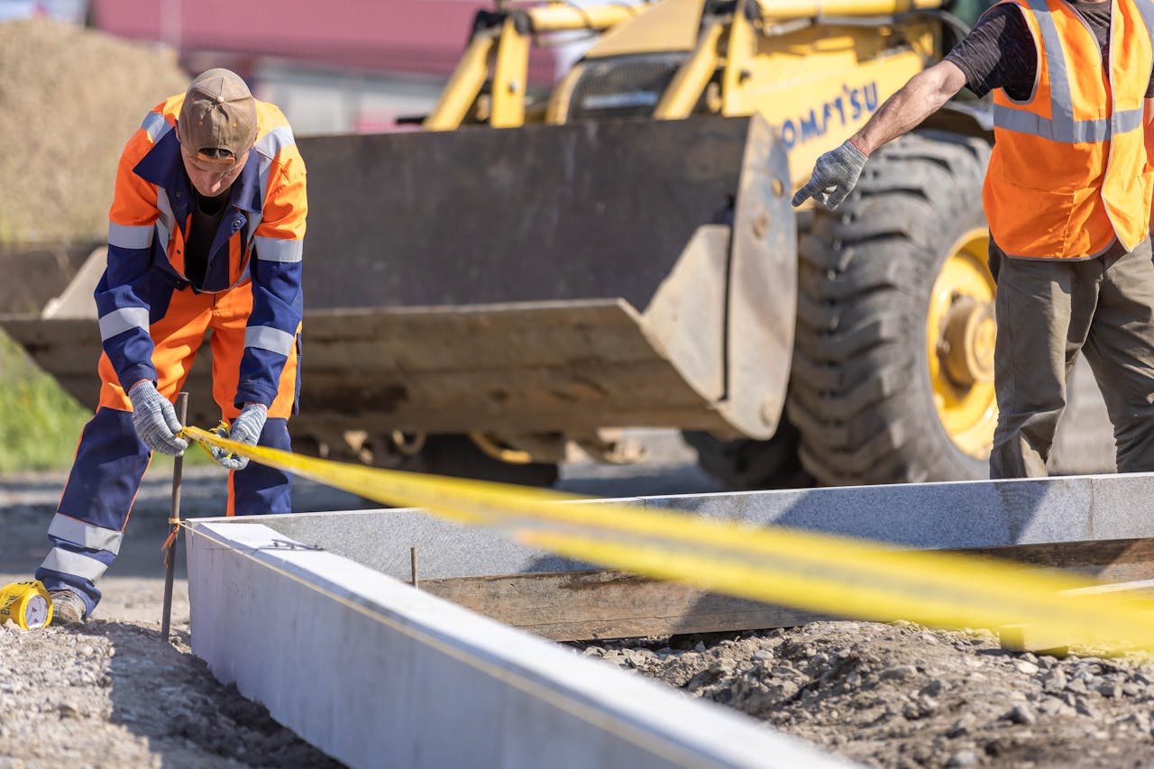 Construction workers using measuring tape at a sunny outdoor site with a bulldozer in the background.